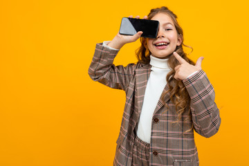 attractive smiling young Caucasian girl in a classic suit shows a phone with a mockup on a yellow background