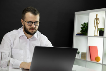 Young handsome bearded freelancer in white shirt and glasses working at laptop, makes video call, communicates with customers and partners. Cozy office workplace, online conference, e learning concept