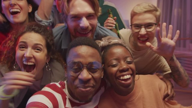 Excited multiethnic friends smiling, waving and posing together for camera while doing group selfie with smartphone at home party