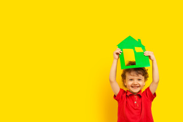 stay home. A boy, a curly laughing emotional kid holds a cardboard house of green color above his head at the top on a bright yellow background, studio. coronavirus pandemic.