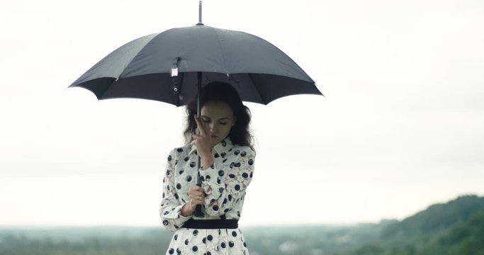 Attractive young elegant girl stands outside in rainy weather with black umbrella. the girl is feeling sad.