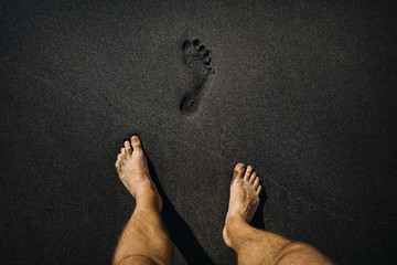 Close up of male footprints and feet walking on the volcanic black sand on the beach - Powered by Adobe