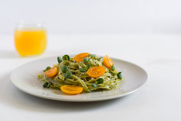 A grey plate of green fresh pasta with olive oil, raw orange tomatoes, micro greens and sesame seeds with orange juice on the background. Horizontal. Copy space on the right.
