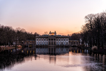 Fototapeta premium Warszawa Lazienki or Royal Baths Park garden with palace building in center by water lake at sunset in Warsaw, Poland at sunset dusk or twilight