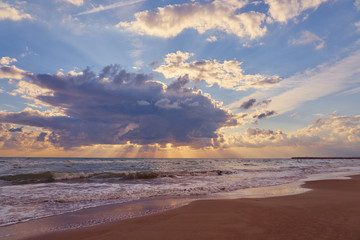 The Sun behind the huge cloud over waving sea and reflections of sky on wet sand on tropical beach.