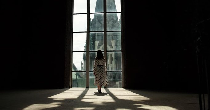 girl in a white dress fits to a large window. the building is visible from the window. girl stands by the window with her hands resting on him.