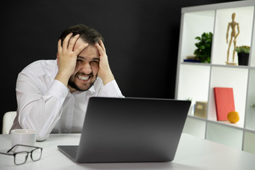 Stressed young man in panic holds hands on head screaming with gritted teeth siitting in front of laptop in home office during quarantine. Business problems, lack of money, crisis, apathy, depression