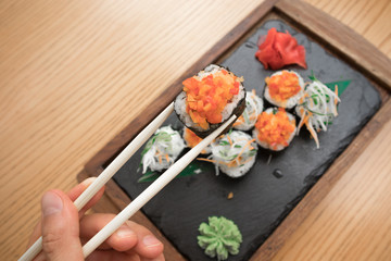close up of a young man's hand holding and eating vegetarian sushi rolls with chopsticks on a wooden tray on a wooden table 