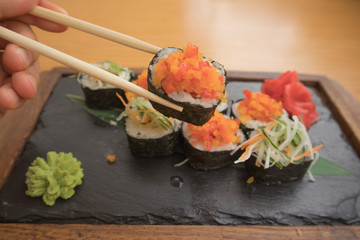 close up of a young man's hand holding and eating vegetarian sushi rolls with chopsticks on a wooden tray on a wooden table 