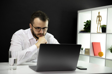Serious attractive bearded mаn in white shirt and glasses working at laptop at home during self-isolation and quarantine. Smart freelancer stares intently at monitor, working from home
