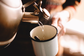 Man pouring boiling water from kettle in mug preparing coffee or tea at home