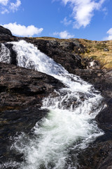 Sunny white scenic norwegian powerful river cascade waterfall with dark rocks and bright blue sky. Nature travel clean falling water vertical landscape