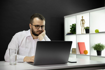 Handsome bearded male freelancer in white shirt and glasses working at computer in home office, tired bored young businessman looking at monitor. Emotional and physical burnout at remote work