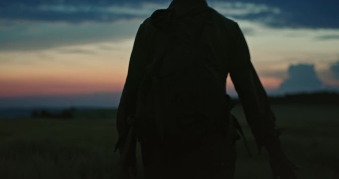Silhouette of serviceman with backpack touching tall grass while walking in lonely remote field with sunset sky. Lonely soldier walking in tall grass of field at sunset.