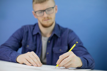 Thoughtful focused young male designer in eyeglass draws with pencil in studio with wooden dummy on blue background. Selective focus on pencil