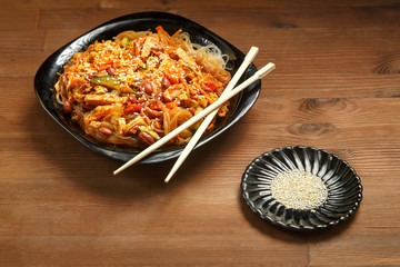 Black plate with funchose and chicken with vegetables under pad thai sauce on a wooden chopstick table.