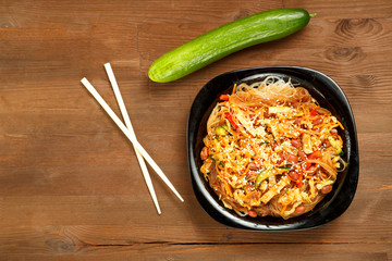 Black plate with funchose and chicken with vegetables on a wooden table and fresh vegetables.