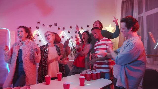 Young African American man drinking alcohol from red cup and showing yes gesture while losing at beer pong game at home party with friends