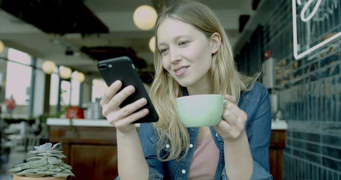 Beautiful Caucasian female looking at smart phone in trendy cafe using smartphone technology and drinking coffee