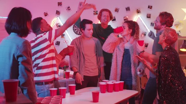 Young joyous woman drinking from red plastic cup while losing beer pong game while excited multiethnic friends cheering for her at home party
