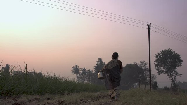 Low angle of Indian woman walking and carrying bucket of water at sunset