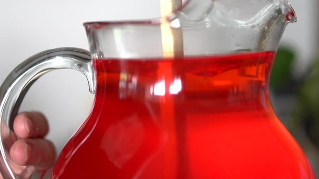 Stirring red drink mix in glass pitcher of water with wooden spoon