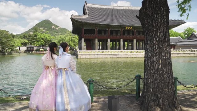 Two girls in traditional Korean hanbok dresses looking at Gyeongbokgung Palace, wide shot truck left in slow motion.