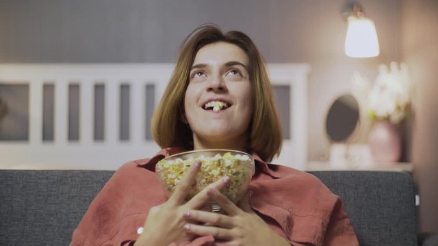 Close up of happy young woman sitting on the sofa, eating popcorn, watching comedy movie and laughing