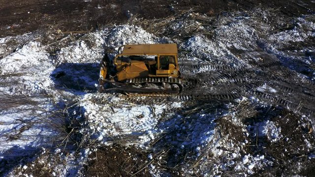 Aerial ascending shot of old yellow bull dozer clearing trees and snow to make new farm land.