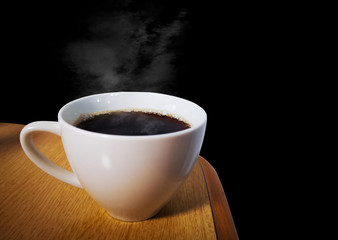 Black coffee with smoke in a white mug placed at the corner of a wooden desk On a black background. Refreshing coffee before working at home.