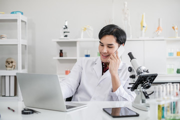 A male scientist with black hair wearing white coat working on laptop and talking on a mobile phone with a microscope in a laboratory setting with test tubes. Looking relax during break time.