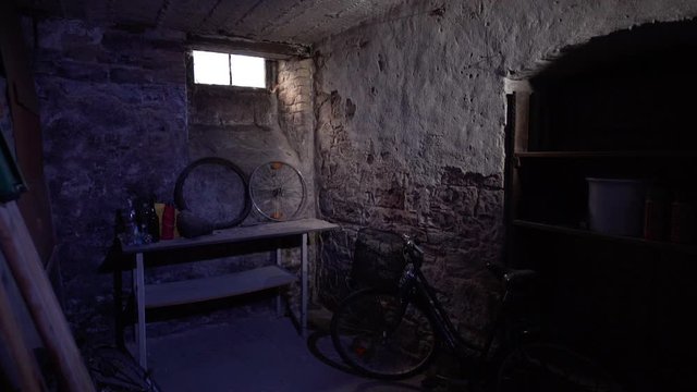 Man enters a stony, dark basement with a workbench and a bicycle. Light enters the room through a small window.