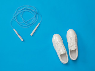 White sneakers and a skipping rope with white handles on a blue background. Sports style. Flat lay.