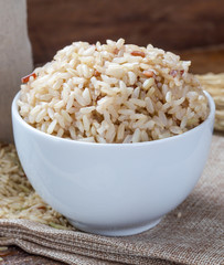 Cook and raw brown rice in white ceramic bowl on wood table and wood background with spoon. Copy space.