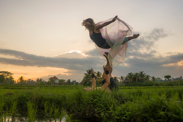 outdoors sunset acroyoga workout - young happy and fit couple practicing acro yoga drill at beautiful rice field enjoying nature doing acrobatic pose with woman in wings