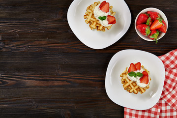 Beautiful breakfast. Belgian Viennese waffles decorated with cream and strawberries,  tea and a napkin on a dark wooden background. Top view.