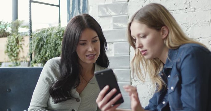 Female friends in cafe looking at smart phone, wireless technology friendship togetherness