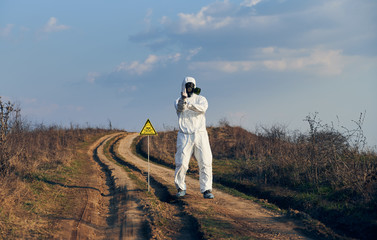 Obraz premium Horizontal snapshot of man in white coverall, gas mask and gloves standing on village road in field next to toxic sign showing middle finger, radiation zone. Concept of ecology, danger and hazard.