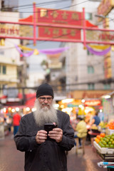 Mature bearded tourist man with eyeglasses using phone in Chinatown