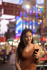 Happy young beautiful Asian tourist woman using phone in Chinatown at night
