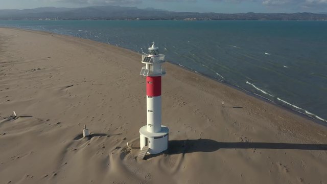 Fangar lighthouse, or delta del ebro, standing alone on wide sandy beach warning ships of navigational hazard where river meets sea in Spain. White and red lighthouse on shore with blue water of sea.