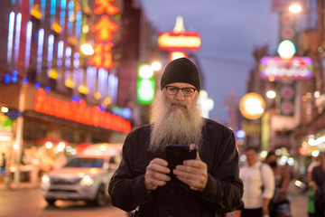 Mature bearded tourist man with eyeglasses using phone in Chinatown at night
