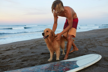 Handsome Surfer. Surfing Man Playing With Dog Near White Surfboard On Sandy Beach. Beautiful...