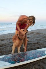 Handsome Surfer. Surfing Man Playing With Dog Near White Surfboard On Sandy Beach. Beautiful Seascape, Waves On Ocean Surface At Tropical Sunset. 