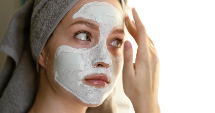 Woman with towel on hairs applying white moisturising mask on face in bathroom, side view. Portrait of young girl making beauty cosmetic treatment fo facial skin care on white background.