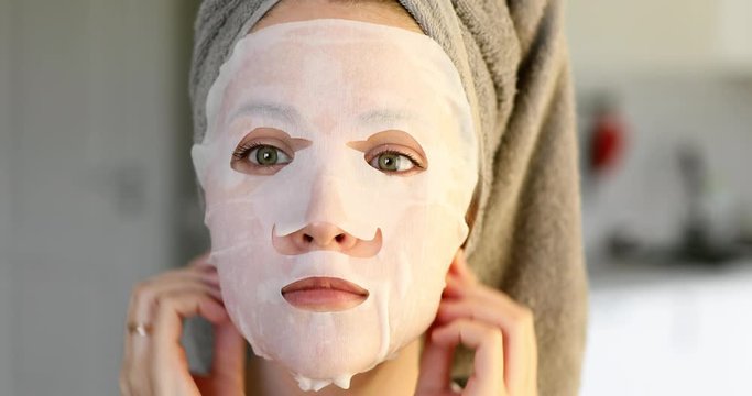 Portrait of woman with towel on hairs applying cosmetic sheet mask on face, front view. Young girl making treatment fo facial skin care. She looking at camera. Beauty treatment. Moisturising of skin.