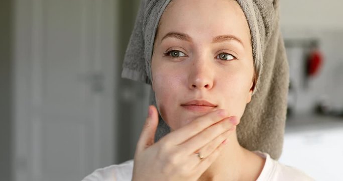 Portrait of young woman with towel on hairs applying cream on face skin, front view. Girl making everyday skincare procedure. Moisturizing and rejuvenation of facial skin. Treatment, cure at home.