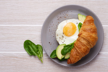 Croissant avocado toast with spinach and fried eggs. Healthy breakfast in plate on light wooden table