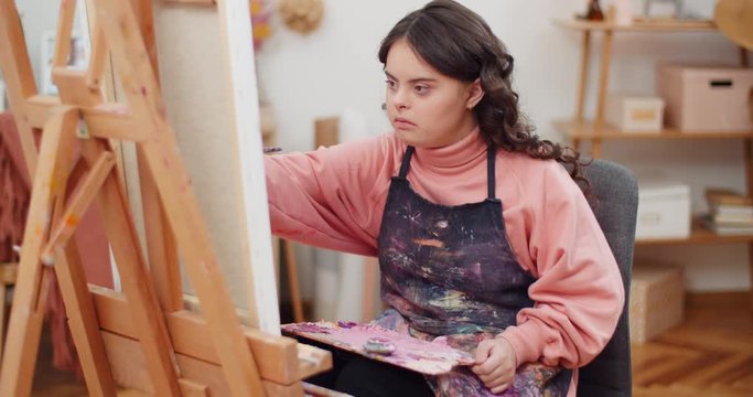 Front view of concentrated beautiful girl with genetic disorder painting in her room. Talanted teenager sitting in front of molbert and holding palette while creating picture.