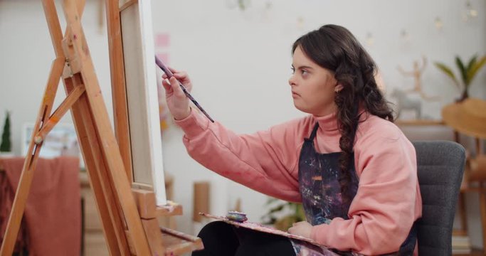 Concentrated schoolgirl in artists apron painting picture while sitting in front of molbert. Pretty teenager with down syndrome sitting with palette on her knees while doing arts. Indoors.